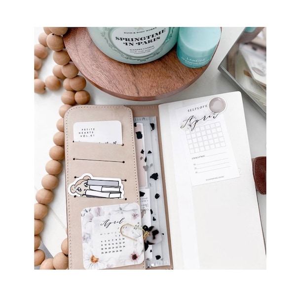 Puede incluir: Una agenda de cuero beige con múltiples bolsillos, que contiene tarjetas y pegatinas. Es visible una pegatina de un oso leyendo libros. También hay un calendario para abril, junto con un desafío de calendario. Una vela de Bath & Body Works y un producto para el cuidado de la piel están en el fondo.