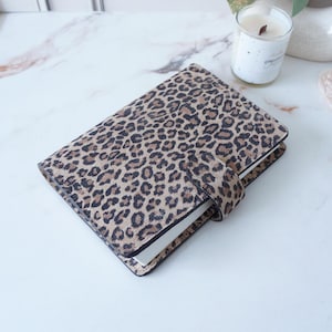 May include: A rectangular planner or notebook cover featuring a brown and black leopard print pattern. The cover includes a matching leopard print strap with a secure closure mechanism, designed to keep the pages protected and neatly contained.