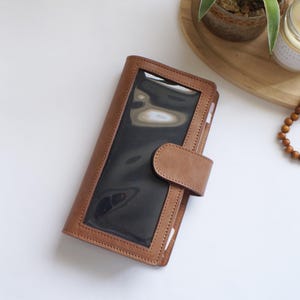 May include: A brown leather wallet with a clear window and a snap closure. The wallet has a rectangular shape and is made of stitched leather. The window is a dark, reflective material. The wallet is lying on a white surface.