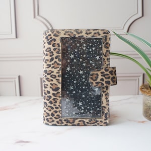 May include: A rectangular journal with a leopard print cover and a clear window showing a starry night design. The journal has a flap closure with a matching leopard print. The background is a light marble surface.