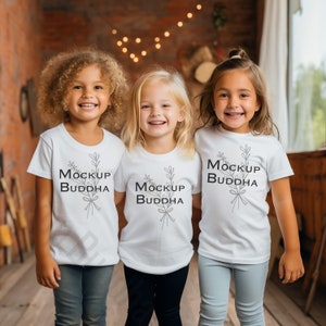 May include: Three children wearing white t-shirts with the text "Mockup Buddha" and a simple floral design. The children are smiling and standing in front of a brick wall.
