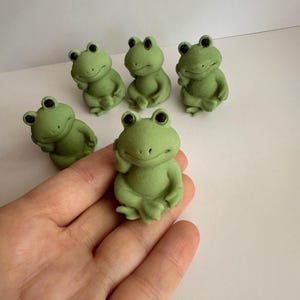 May include: Five small, light green frog figurines with smooth, matte textures and black eyes. Each frog is depicted in a seated, thoughtful pose with one hand resting near its chin. The figures are uniform in colour and design, showing a consistent shape.