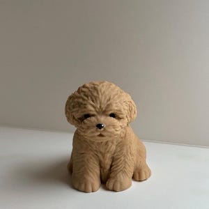 Könnte beinhalten: Eine kleine, hellbraune Figur in Form eines sitzenden Welpen mit lockigem, strukturiertem Fell. Der Welpe hat einen runden Kopf, Schlappohren und eine kleine schwarze Nase. Die Skulptur besteht aus einem soliden, matten Material mit Details.
