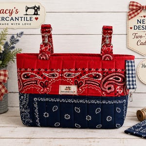 May include: A quilted two-tone caddy featuring a red upper section with a white and black paisley bandana print and a navy blue lower section with a white floral bandana pattern. It has two fabric handles attached with silver metal rings.