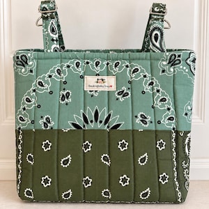 May include: A quilted tote bag featuring a two-tone green bandana print with black and white paisley patterns. The bag has vertical stitching, two fabric straps with silver-toned metal hardware, and a central label reading Handcraftedby Tracy.