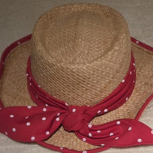 Puede incluir: Un sombrero de paja trenzada de color tostado con ala ancha y copa redondeada. Una cinta de tela roja con lunares blancos está atada alrededor de la base de la copa, con los extremos anudados al frente. El borde del ala tiene un ribete de tela roja.
