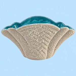 May include: A scalloped ceramic bowl featuring a textured, sandy-beige exterior finish and a smooth, glossy teal interior. The bowl has a wide, open design with a curved rim, showcasing a distinct contrast between the rough outer surface and the vibrant, polished blue-green glaze inside the vessel.