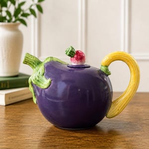 Peut inclure: Une théière en céramique en forme d'aubergine violette avec un bec verseur en forme de tige verte, une anse jaune incurvée et un couvercle surmonté d'un petit radis. La théière présente une finition brillante et des textures réalistes.