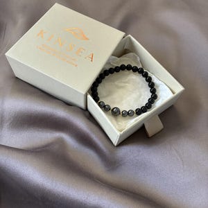 May include: A beaded bracelet featuring smooth black spherical beads and a central cluster of metallic grey beads, presented in a beige cardboard box. The box lid is printed with the gold text KINSEA and a leaf icon above smaller text.