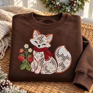 May include: A dark brown crewneck sweatshirt featuring an embroidered design of a white lace-patterned fox wearing a red scarf, positioned next to a small green plant with three red strawberries and white blossoms on the front.