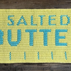 May include: A rectangular crochet mat in light yellow with the text "SALTED BUTTER" in teal block letters. The mat features a series of vertical teal lines along the bottom edge and has short, fringed teal and yellow yarn edges on both sides.
