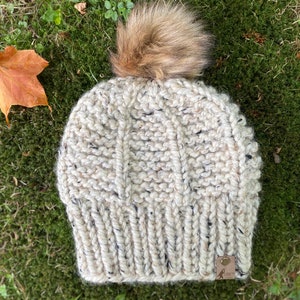 Puede incluir: Un gorro de punto blanco y negro moteado con un pompón marrón esponjoso en la parte superior. El gorro tiene una pequeña etiqueta de cuero marrón con el texto "LOFT" en ella.