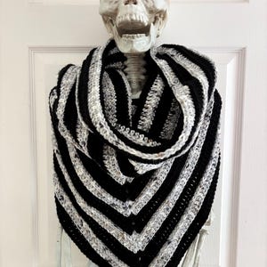 May include: A triangular crochet shawl featuring alternating stripes of solid black yarn and a variegated grey, white, and black speckled yarn. The shawl has a V-shaped point at the bottom and is draped around the neck of a human skull model.
