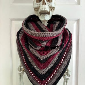 May include: A crochet cowl scarf featuring a V-shaped design with alternating horizontal stripes in black, deep burgundy, and muted grey. The textured yarn accessory displays a repeating pattern of solid stitches and openwork rows, forming a triangular drape.