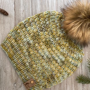 May include: A green, yellow, and white knit beanie with a brown faux fur pom pom. The beanie has a brown leather tag with the text "Looney Lass" on it.