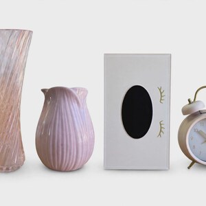 Peut inclure: Un grand vase en verre rose translucide avec un motif en spirale, un vase en céramique rose opaque plus court avec des stries verticales, une boîte à mouchoirs rectangulaire blanche avec une ouverture ovale noire et des cils dorés, et un réveil rose.
