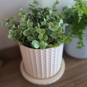 May include: A light pink cylindrical plant pot featuring a geometric diamond-patterned texture on its exterior surface. The pot sits on a matching circular saucer and contains a lush, green artificial plant with small, rounded leaves.