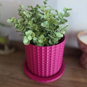 May include: A vibrant magenta cylindrical plant pot featuring a geometric diamond-patterned texture on its exterior. The pot includes a matching circular saucer base and contains a small, bushy artificial plant with light green rounded leaves.