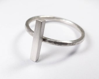 May include: A silver ring with a hammered band and a vertical bar design. The bar is attached to the ring and extends upwards.
