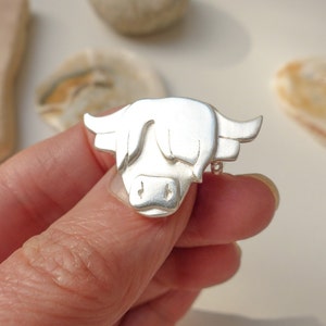 May include: A silver Highland cow brooch, a traditional Scottish symbol, is shown in a close-up view. The brooch is detailed with a textured, three-dimensional design.