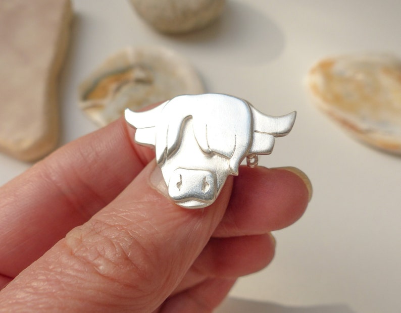 May include: A silver Highland cow brooch, a traditional Scottish symbol, is shown in a close-up view. The brooch is detailed with a textured, three-dimensional design.