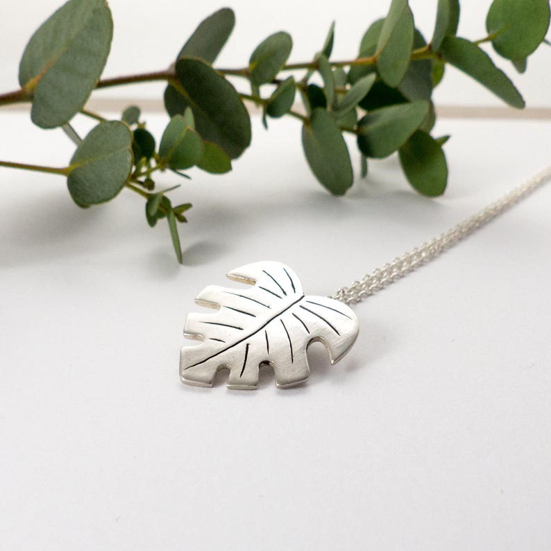 May include: A silver necklace with a detailed monstera leaf pendant. The pendant is textured and has a realistic leaf shape.