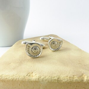 May include: A pair of silver cufflinks with a spiral shell design. The cufflinks are on a light brown surface.