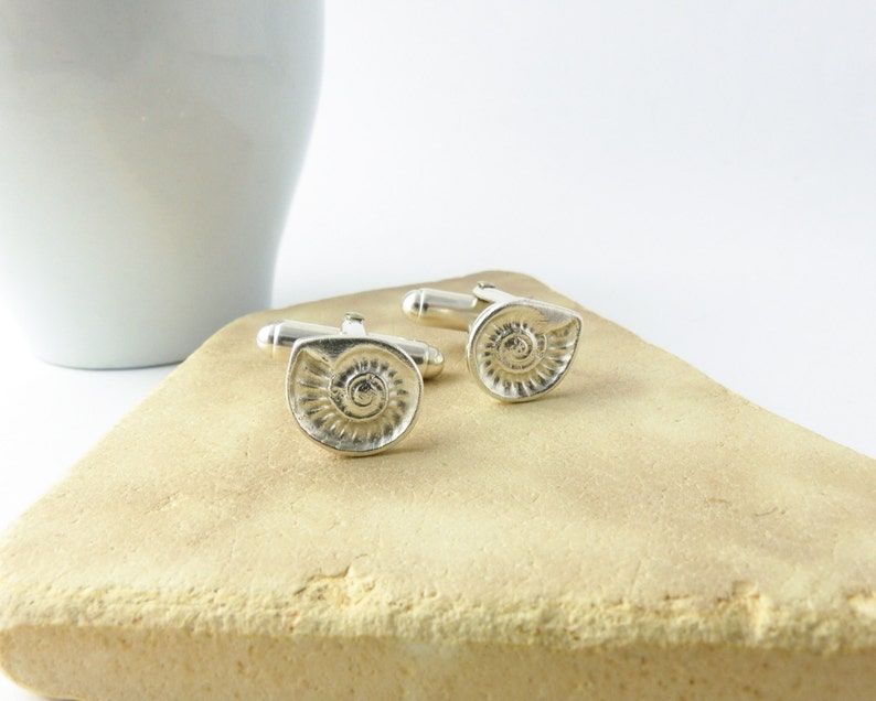 May include: A pair of silver cufflinks with a spiral shell design. The cufflinks are on a light brown surface.