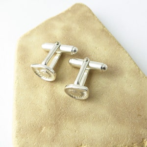 May include: A pair of silver cufflinks with a textured, circular design. The cufflinks are on a light brown surface.
