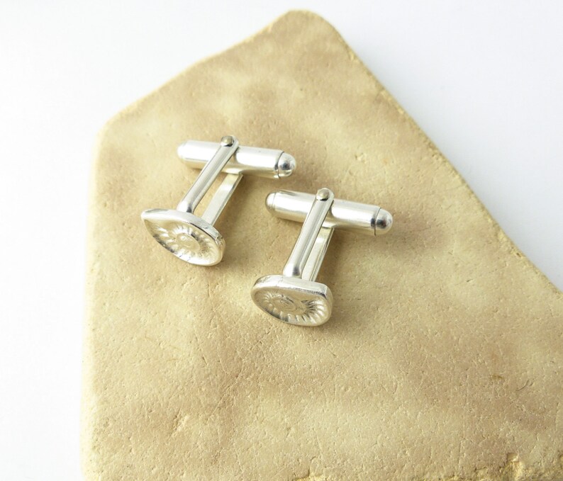 May include: A pair of silver cufflinks with a textured, circular design. The cufflinks are on a light brown surface.