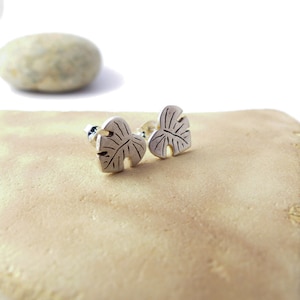 May include: A pair of silver earrings shaped like monstera leaves. The earrings are on a light brown surface.