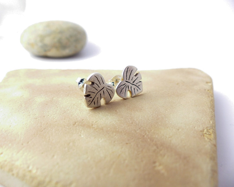 May include: A pair of silver earrings shaped like monstera leaves. The earrings are on a light brown surface.