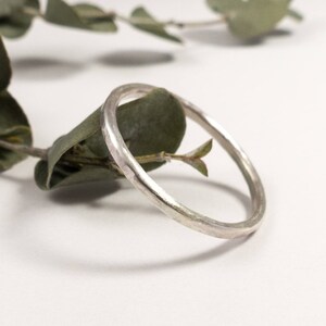 May include: A simple silver band ring with a hammered texture. The ring is shown on a white background with green leaves.