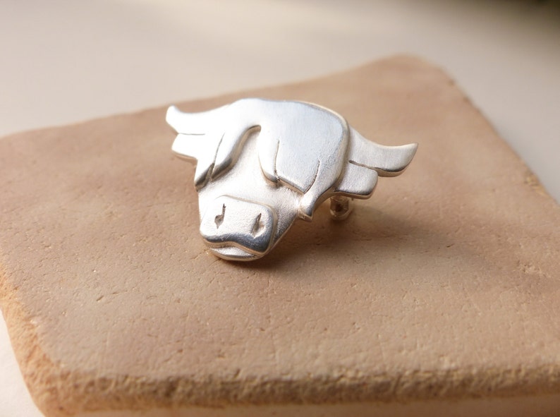 May include: A silver Highland cow brooch, a traditional Scottish symbol, with a detailed design and a brushed finish.