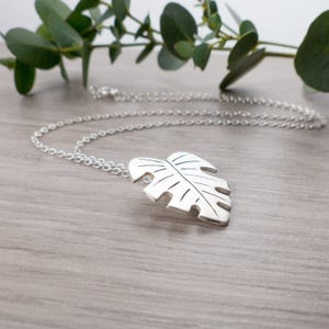 May include: A silver necklace with a large, detailed monstera leaf pendant. The necklace is on a wooden surface with a sprig of greenery in the background.