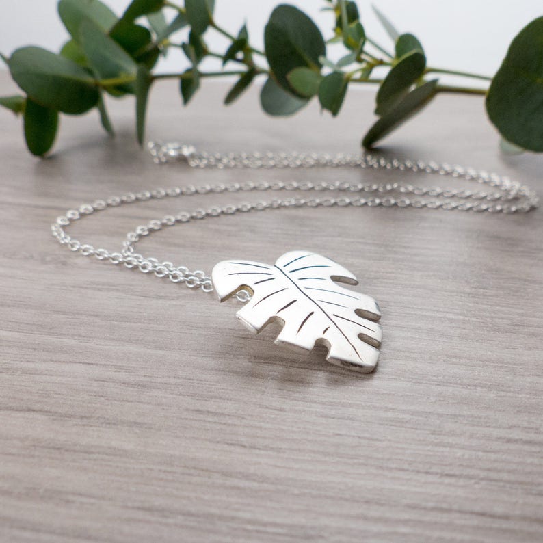 May include: A silver necklace with a large, detailed monstera leaf pendant. The necklace is on a wooden surface with a sprig of greenery in the background.