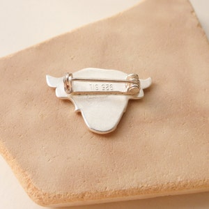 May include: Silver-toned brooch in the shape of a cow's head with a pin on the back. The back of the brooch is marked with "925" and "TIS".