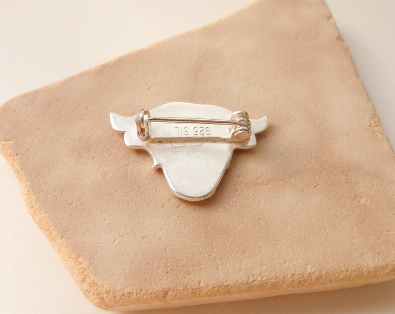 May include: Silver-toned brooch in the shape of a cow's head with a pin on the back. The back of the brooch is marked with "925" and "TIS".
