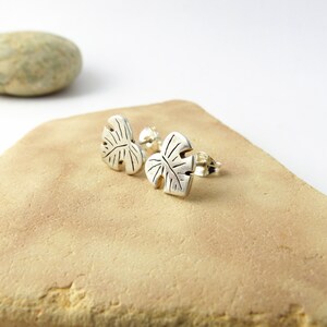 May include: A pair of silver earrings shaped like monstera leaves. The earrings are on a light brown surface.