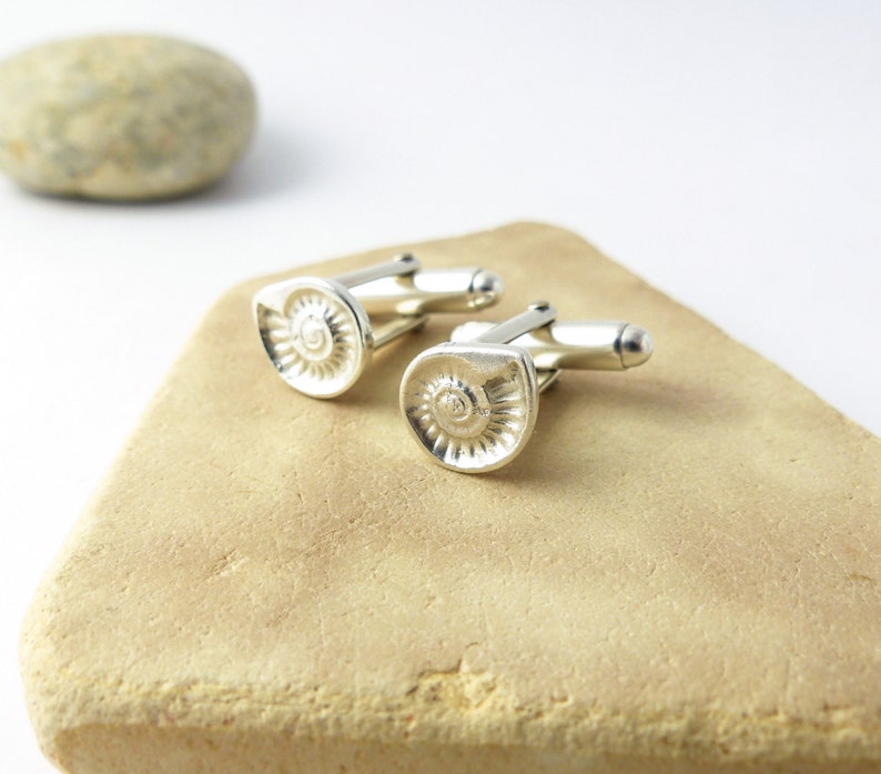 May include: A pair of silver cufflinks with a detailed spiral design. The cufflinks are displayed on a light brown surface.