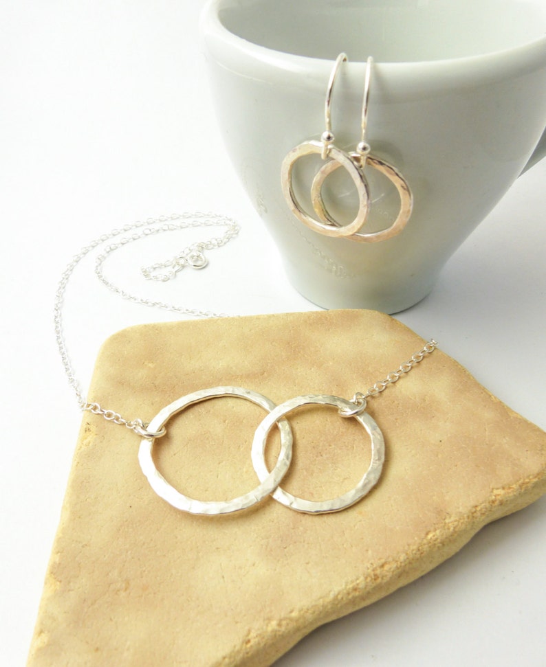 May include: A silver necklace with two interlocking hammered circles. The necklace is on a brown surface. A pair of silver hammered circle earrings are in a white cup.
