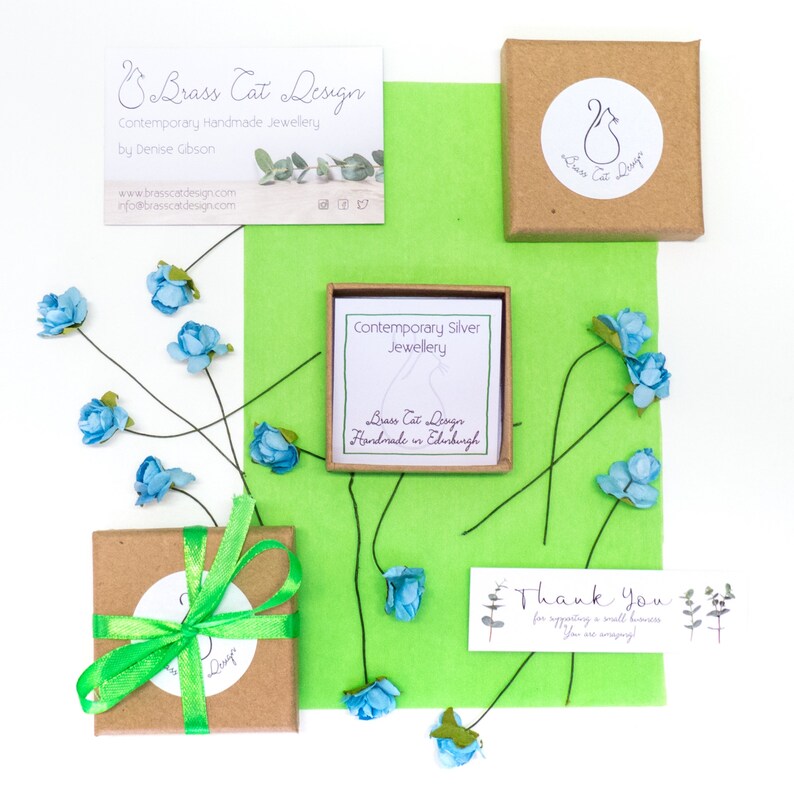 May include: A small brown box with a green ribbon and a thank you card with the text "Thank You for supporting a small business. You are amazing!"  The box is surrounded by blue flowers and a green background. The box is labeled "Brass Cat Design Handmade in Edinburgh".
