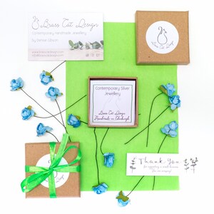 May include: A small brown box with a green ribbon tied around it. The box is sitting on a green surface with blue flowers. The box has a white label that says "Brass Cat Design" and "Handmade in Edinburgh". There is a thank you card on the surface that says "Thank You for supporting a small business. You are amazing!"