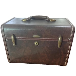 May include: A dark brown rectangular vanity case with a textured finish, featuring a top handle, brass-toned metal latches, and a central keyhole. The edges are accented with white stitching, and a small oval metal brand plate is centered on the front.