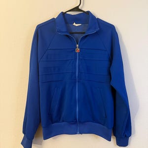 May include: A vibrant royal blue track jacket featuring a full-length front zipper with a silver-toned metal pull tab displaying a red and white letter B logo. The jacket includes a high collar, long sleeves with ribbed cuffs, and horizontal pleats.