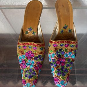 May include: A pair of tan-colored, open-back mules with intricate floral embroidery in shades of pink, blue, purple, and green. The heels have a tan leather insole with a floral design. The shoes are likely vintage or handmade.
