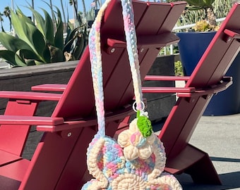 May include: A crochet flower-shaped bag featuring a pastel variegated yarn in shades of pink, blue, and yellow with a cream-colored border. The bag includes a long matching strap and a small, separate crochet flower charm attached to the top.