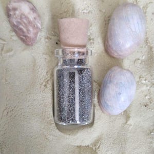 May include: A small clear glass bottle with a cork stopper filled with fine dark grey sand. The bottle is surrounded by three smooth, oval-shaped seashells in shades of light brown, beige, and pale grey, all resting on a bed of light sand.