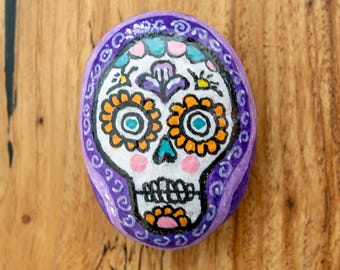 May include: A hand-painted rock featuring a white skull with black outlines, orange flower eye sockets, a teal nose, and pink cheeks. The skull is framed by a purple border decorated with white swirls and colorful floral accents.