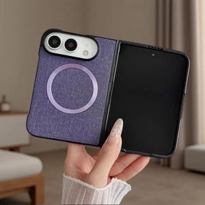 May include: A foldable smartphone case with a textured purple fabric finish. The back features a circular magnetic ring and a black camera housing with two lenses and a flash. The front displays a black screen with a small circular camera cutout.